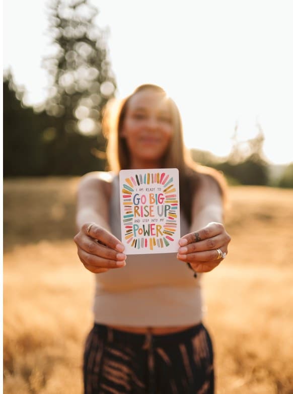Benisha in a sunlit golden field holding out an affirmation card that reads: I am ready to go big, rise up and step into my power